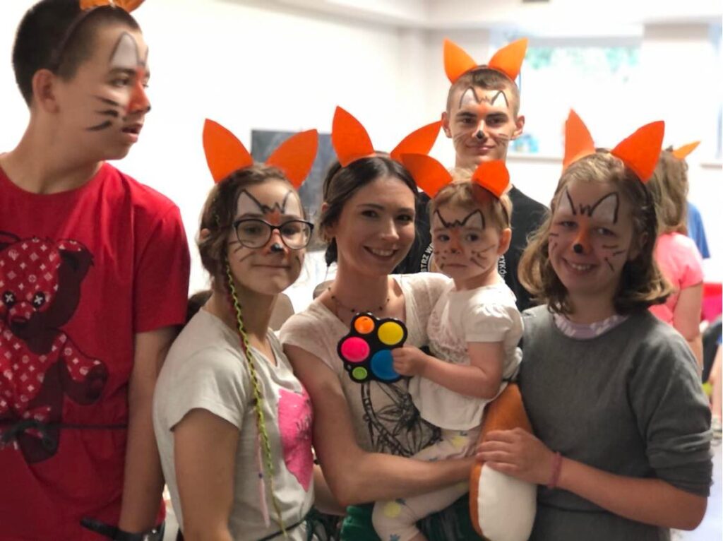 Uśmiechnięta grupa patrzy w obiektyw. Czwaoro dzieci, a między nimi kobieta z rocznym dzieckiem na rękach. Wszyscy mają pomarańczowe uszy na głowie i pomalowane twarze - wyglądają jak liski.